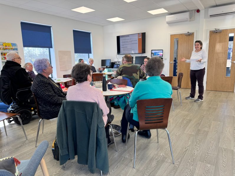 Littlemore Flowers of Proximity workshop 2025 A person presents to a seated audience in a bright community room.